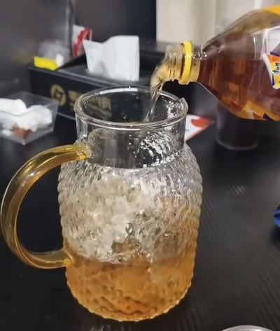 办公室桌面上，一只透明玻璃水壶正在被倒入棕色液体（疑似茶饮料）。水壶表面有凸起纹理设计，搭配金色把手，液体倒入时产生细密气泡。背景可见电脑显示器、纸巾盒和办公杂物，整体呈现日常办公场景的一角。