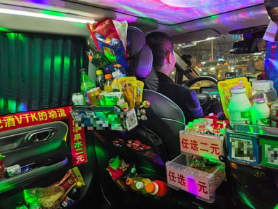夜晚的出租车内，车顶布满彩色LED氛围灯，营造出梦幻的星空效果。车内后排被各种零食饮料填满，包括瓶装水、袋装零食等，座椅靠背上挂满了商品，旁边还有标着“任选二元”的塑料收纳盒。车窗挂着绿色窗帘，车门上贴着红色标语，整体看起来像一个移动的小型便利店。司机正专注地驾驶车辆，前方可以看到城市夜晚的街景和车流。