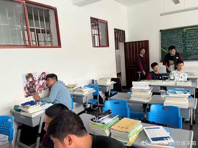 一间典型的中学教室场景，课桌上堆满了厚厚的书本和试卷，蓝色的塑料座椅整齐排列。一名穿浅蓝色连帽衫的男生正站在教室左侧墙边，专注地将一张印有二次元美少女角色的海报贴在墙上，海报上的角色有着粉色头发和精致的服饰。教室后方有几名男生或站或坐，目光似乎投向贴海报的同学，黑板上写满了绿色粉笔的板书，内容涉及学习方法或班级规定。整体氛围轻松真实，展现了校园生活中略带调皮的日常瞬间。