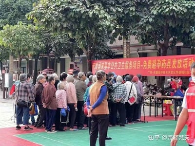 这是广西-东盟经济技术开发区华侨城社区的户外公益活动现场，一条红色横幅标明活动为「开展国家基本公共卫生服务项目-免费避孕药具发放活动」。大批老年居民聚集排队，等待领取相关物资，穿红马甲的工作人员在服务台前忙碌准备。背景有茂盛的树木和停放的电动车，场地是红绿相间的户外防滑地面，整体呈现出社区公共卫生服务活动的热闹场景。图中没有双关、谐音梗或暗示内容。