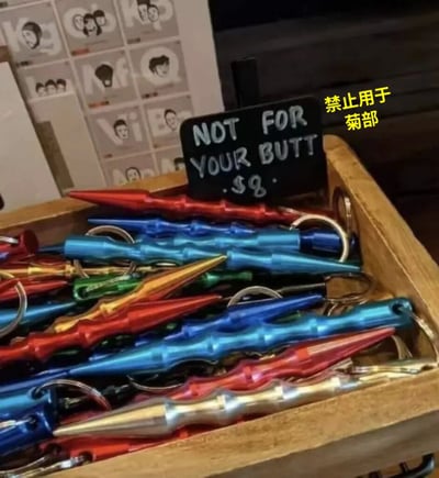 图片展示了一个木质 crate 里装满了彩色（红、蓝、绿、金等）尖刺状钥匙扣，上方放置着一块黑色小标牌，用白色字体写着“NOT FOR YOUR BUTT .$8”。右侧有黄色中文文字叠加“禁止用于菊部”（“菊部”为网络委婉语，指代肛门部位）。背景中可见模糊的人物头像海报。图片通过物品形状与警示文字的反差制造幽默效果，暗示该尖刺钥匙扣可能被误用的场景。