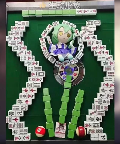 这是一张在绿色麻将桌上的创意摆拍作品，玩家用各类麻将牌（万子、筒子、条子、风牌、箭牌等）摆出了一对展开的翅膀造型，翅膀中间放置了一个初音未来的毛绒玩偶，下方用绿色麻将牌摆出装饰结构，还搭配了两个红白相间的球和一张小卡片，麻将桌的中心转盘也作为背景的一部分。图片顶部有文字“生动形象”，将二次元玩偶与麻将结合，充满趣味性和创意，属于玩家的趣味脑洞创作。