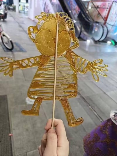 图片近距离展示了一只手握着的金色糖人，糖人呈卡通人物造型，有着圆圆的脸庞、大大的笑脸、卷曲的头发细节，双臂向两侧张开，身穿条纹图案服饰，整体由透明金黄的糖料制成，纹路清晰精致。背景为繁忙的城市街头或市场环境，可见自动扶梯、停放的自行车及模糊的店铺招牌，展现了传统手工艺在现代都市场景中的呈现。