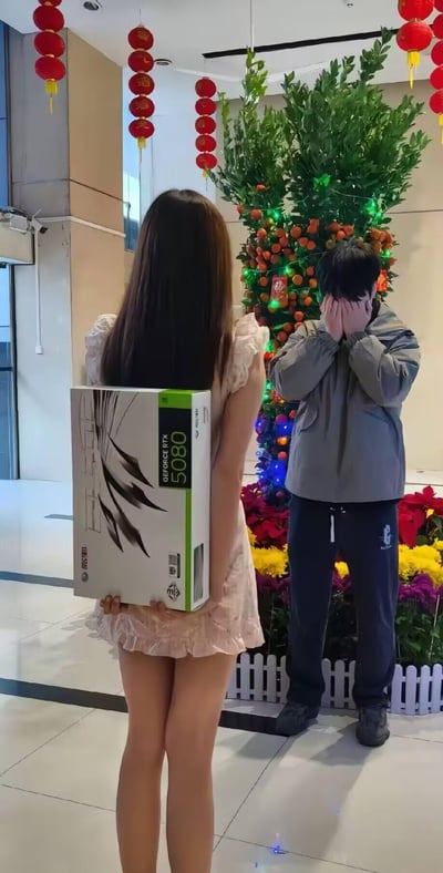 在充满春节氛围的室内场景中，悬挂着印有“酒酒”字样的红色灯笼，一旁还有装饰着彩色灯带与年桔的绿植。一位长发女生背对着镜头，将NVIDIA GeForce RTX 5080显卡的包装盒藏在身后，似乎要给面前的男生送上惊喜；对面的男生则用双手紧紧捂住脸部，看起来既害羞又激动，显然被这份投其所好的礼物触动。