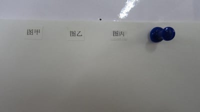 这是一张横向的原始拍摄照片，画面主体为一张浅色空白纸张，纸张上方从左至右依次粘贴着“图甲”“图乙”“图丙”三个白色小标签，纸张右侧固定着一个蓝色的双头按钉，纸张上方露出一小部分白色板面。整体画面简洁，仅用于标记后续需补充的图甲、图乙、图丙内容，无特殊双关、谐音梗或暗示含义。