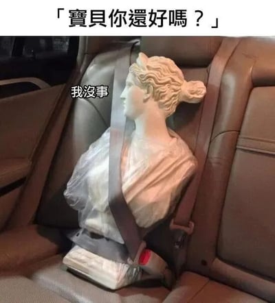 图片展示了一尊古典风格的大理石胸像被安全带固定在汽车的乘客座椅上，雕像神态平静，发丝和衣纹细节清晰。图片上方配有文字「「寶貝你還好嗎？」」，仿佛有人在关切询问，而雕像旁边则出现「我沒事」的回应文字。通过将庄重的古典雕塑置于日常汽车场景中，并赋予其拟人化对话，形成了强烈的反差萌，营造出幽默搞笑的效果。