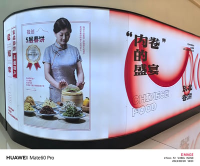 这是一张商场或餐厅内的大型弧形广告海报，左侧展示一位身着浅蓝色旗袍的女性，双手捧着刚制作好的5层春饼，背景配有‘独创5层春饼’‘非物质文化遗产’等文字及多盘配菜（如肉丝、豆芽、黄瓜丝），右侧以红色渐变背景突出‘内卷的盛宴’黑色大字，下方标注‘CHINESE FOOD’及‘东北卷饼’。海报巧妙运用网络热词‘内卷’与‘卷饼’的‘卷’形成双关，既贴合当下语境又突出产品特性。底部有水印‘HUAWEI Mate60 Pro’‘X-MAGE’及拍摄时间‘2024/08/28 14:03’。