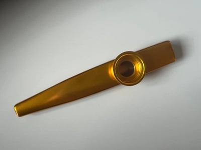 一张金色滑笛乐器的静物照片，主体为黄铜色长条形物体，中间有圆形吹口，整体呈现光滑金属质感。物体放置在纯白色背景上，通过柔和的侧光形成轻微阴影，突出器物的立体感和金属反光特性。