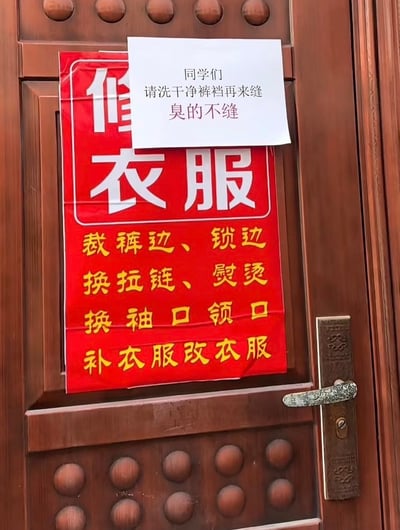 图片展示了一扇木质门上贴着的两张告示。下方是红色底白字的“修衣服”招牌，用黄色字体列出了“裁裤边、锁边、换拉链、熨烫、换袖口领口、补衣服改衣服”等服务项目。招牌上方叠加了一张白色手写便签，内容为“同学们 请洗干净裤裆再来缝 臭的不缝”，直白又幽默的提示让人会心一笑，推测是面向学生群体的修衣店老板为避免尴尬而贴出的“特殊要求”。