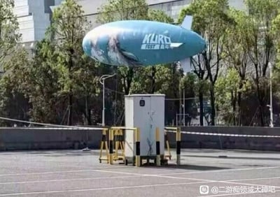 这是一张户外拍摄的视觉错位趣味图片，画面中印有“KURO FEST”字样以及库洛游戏角色图案的浅蓝色宣传飞艇正悬浮在空中，从拍摄者的角度看，飞艇刚好位于地面上的白色移动公厕的正上方，二者位置巧合形成了仿佛飞艇是公厕顶盖、二者为一体装置的搞笑错觉。背景可见成片的行道树与远处的城市建筑，地面是空旷的铺装场地，图片右下角带有百度贴吧“二游痴领域大神吧”的水印。