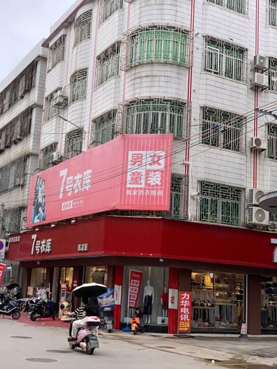 这是一张充满生活气息的城镇街头实拍图，画面核心是一家名为「7号衣库（陈店站）」的服装店铺，店铺采用醒目的红色门头与大型红色广告牌，广告牌上标注着「男女童装 我家的衣帽间」的宣传语。店面前方，一名撑着黑伞的路人正骑着粉色电动车经过，背景是带有绿色防盗窗与多台空调外机的居民楼，空中交错着电线，整体呈现出平实的城镇日常风貌。