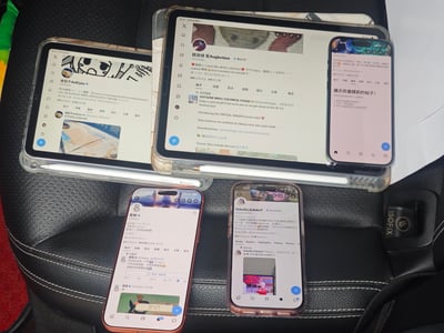 这是一张实拍照片，在汽车的黑色真皮座椅上，整齐摆放了两台平板电脑和三台智能手机，共五台电子设备，每台设备的屏幕都显示着X（原推特）社交平台不同二次元创作者的个人账号主页，是爱好者线下碰面时集体展示账号的场景。