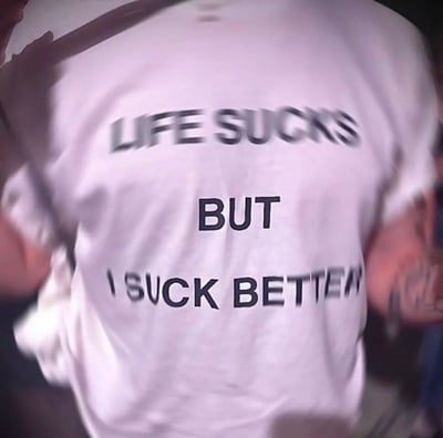 图片展示了一个人穿着白色T恤的背影，T恤上印有黑色英文标语："LIFE SUCKS BUT I SUCK BETTER"。这句话以自嘲的方式表达了尽管生活不如意，但自己比生活更会