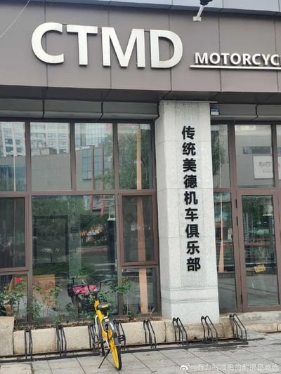 图片展示了一家临街机车俱乐部的外观。门头上方醒目地标注着大写英文字母“CTMD”及“MOTORCYC”字样，门柱上则竖排刻有“传统美德机车俱乐部”的中文招牌。店面前的停放架上，一辆黄色共享单车斜倚着，玻璃橱窗清晰倒映出周边的高楼建筑。画面中存在明显的双关设计：“CTMD”虽是“传统美德”的拼音首字母缩写，但在网络语境中常被用作谐音粗话，这种严肃命名与俚语的碰撞形成了强烈的幽默反差。