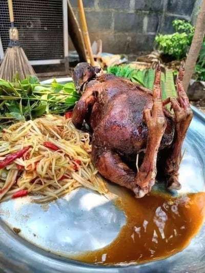 一张展示传统美食的照片，画面中央是一只烤得焦香的整鸡，呈现出诱人的深棕色，鸡皮酥脆。烤鸡被放置在一个金属大盘中，周围配有新鲜的绿色蔬菜、香菜和一盘凉拌姜丝，姜丝中还点缀着红色辣椒丝增添色彩。背景中可以看到一些家庭用品和绿植，营造出温馨的家常烹饪氛围。