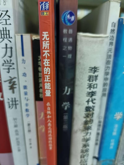 图片展示了书架上一排书籍的特写，主要为力学、数学等领域的学术著作，如《经典力学辛讲》《力、功、能量与辛数学》《力学（第三版）》《李群和李代数对约束力系统的应用》等。其中一本红色封面的书籍《无所不在的正能量 在自然和人类之间流淌的故事》格外显眼，与周围严肃的学术书籍形成鲜明对比，营造出一种幽默的反差感。
