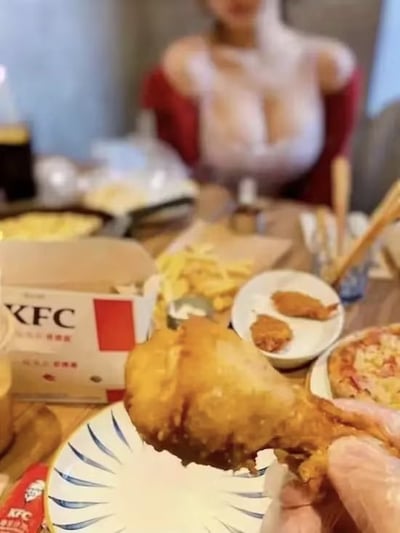 图片近距离呈现了一只金黄酥脆的KFC炸鸡腿被手持着，外皮色泽诱人，质感饱满。前景清晰聚焦于鸡腿，背景为摆满KFC餐点的餐桌，可见薯条、KFC餐盒等食物，桌边坐着一位穿着红色上衣的女性，背景略微虚化以突出前景美食。整体场景营造出享受KFC美食的愉悦氛围，传递出食物美味的直观感受。