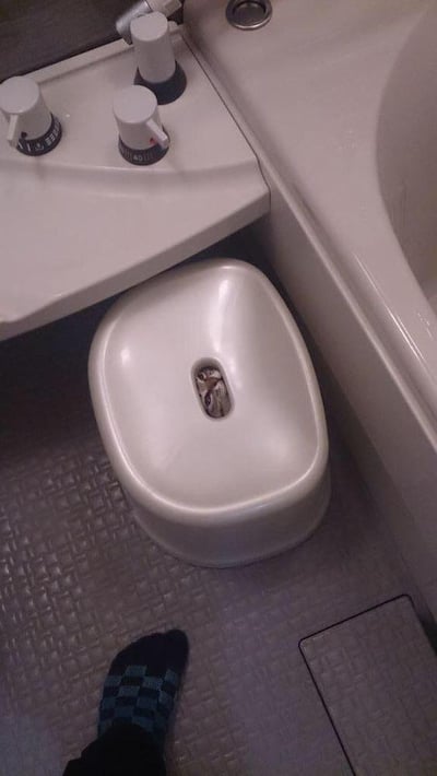 这是一张在浴室场景下拍摄的趣味视觉错位照片。画面中央的坐浴盆（净身盆）排水口位置，恰好与一只猫头鹰的头部完美重合，营造出猫头鹰“待在”洁具内部的错觉。画面左上角可见浴缸的水温控制旋钮，右侧是米白色的浴缸边缘，左下角能看到一只穿着蓝黑格子防滑袜子的脚，地面铺设着带有纹理的防滑浴室地砖。猫头鹰的头部从洁具洞口露出，表情看起来有些茫然，巧合的位置搭配让整个场景显得十分滑稽有趣，属于典型的生活中偶然出现的视觉错位趣味场景。