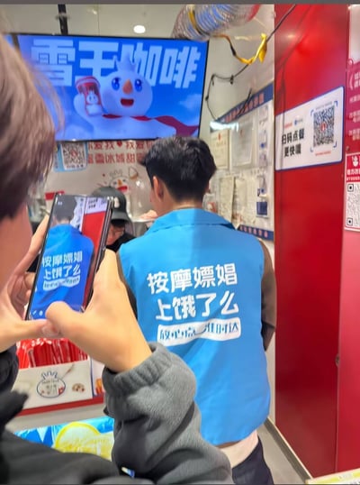 图片显示一家咖啡店内部场景，店内墙上挂着显示'雪王咖啡'字样和卡通雪人头形象的屏幕。一名身穿蓝色背心的工作人员背影面向柜台，其背心上印有争议性文字'按摩嫖娼 上饿了么 做中点工作团队'。另一个人正拿着手机对着这件背心拍照。店内还可见到一些宣传海报和二维码。