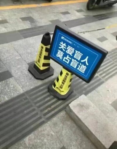 这张实拍照片展现了一个极具讽刺性的城市场景：街道的盲道上被两个黄黑相间的路锥占据，其中一个路锥上还立着一块蓝色提示牌，上面清晰写着“关爱盲人 莫占盲道”的公益标语。标语倡导的内容和实际占用盲道的行为完全相悖，形成强烈反差，讽刺了现实中部分场景里，无障碍设施被随意占用、公益口号流于表面的现象，引人反思对无障碍环境的重视程度。