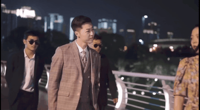 图片展示了《龙王赘婿》广告系列的经典场景。夜晚的城市背景下，管云鹏饰演的主角身着格子西装，内搭马甲领带，带领两名黑衣墨镜保镖前行，前方有一位穿着花裙子的女性正面对他们。这一场景体现了“卑微赘婿-身份揭露-打脸逆袭”固定模式中的身份转变时刻，主角展现出与此前“卑微”形象截然不同的霸气冷峻气场。