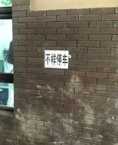 一张拍摄于日常环境中砖墙的照片，墙上固定着一个白色警示牌，牌上用黑色马克笔写着“不样停车”四个大字，明显是“不准停车”的错别字。左侧有一扇玻璃窗，窗内隐约可见模糊的人影或物品。整体场景因错别字产生了幽默效果，展现了生活中常见的文字使用错误现象。