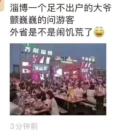图片上方为文字内容：“淄博一个足不出户的大爷颤巍巍的问游客 外省是不是闹饥荒了😆”，下方是一张淄博夜市的实拍照片。照片中夜晚的淄博夜市人头攒动，大量游客聚集在户外餐桌旁用餐，周围霓虹招牌林立，包括“齐聚淄博”“干杯世界”等字样，场面十分热闹。图片通过幽默的文字与拥挤的夜市场景形成反差，调侃淄博旅游热度之高，以至于让不常出门的大爷误以为外省游客因“饥荒”而来淄博觅食，充满诙谐趣味。