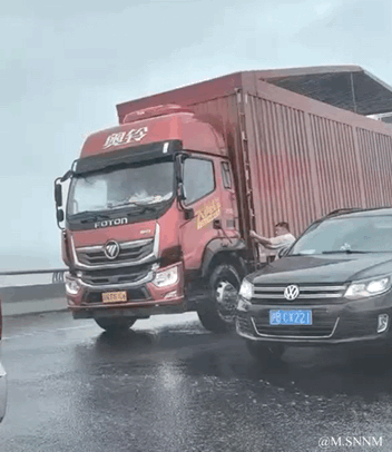 这是一段GIF动态图，呈现了雨天道路上的有趣视觉错位场景：一辆红色奥铃大货车与黑色大众轿车并排行驶，大货车侧厢门开启，一名人员从车厢内探出身，因两车位置重叠，视觉上仿佛是该人员在拍打黑色轿车的车窗，形成了极具迷惑性的趣味错觉，让人第一眼容易产生误解。