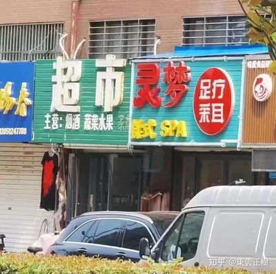 这是一张实拍的街边商铺图，绿色的超市招牌与右侧的灵梦足疗采耳店招牌紧密相邻，视觉上很容易被误读成“超市灵梦”，形成了有趣的巧合。图中左侧超市主营烟酒、蔬菜水果；右侧店铺提供足疗、采耳、泰式SPA服务，画面还包含其他商铺招牌，下方停放着汽车和货车，是日常的城市街边商铺场景。