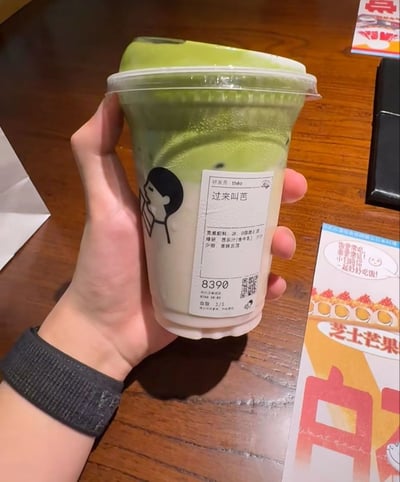 这是一张消费者实拍的图片，画面中一只佩戴黑色织物腕带的手握着一杯喜茶饮品，饮品上层是浅绿色的冰沙/奶盖质地，下层为乳白的牛乳基底，杯身贴的订单标签上饮品名称写为“过来叫芭”，利用饮品配料中的芭乐汁的“芭”和“爸”的谐音制造搞笑效果，背景为木质桌面，右下角露出部分餐饮品牌的芝士芒果相关宣传物料。