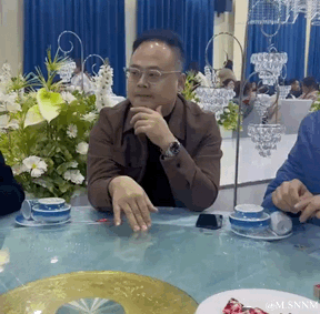这是一段宴会场景的动态画面，画面中心是一位戴眼镜的中年男子，他坐在摆放着蓝白餐具和装饰花的餐桌前，一只手托着下巴，另一只手在桌面上来回滑动，神情看起来若有所思。背景是布置好的宴会场地，能看到其他宾客和精致的水晶吊灯，整体氛围是正式的宴席场合。图中没有双关、谐音梗或暗示内容。