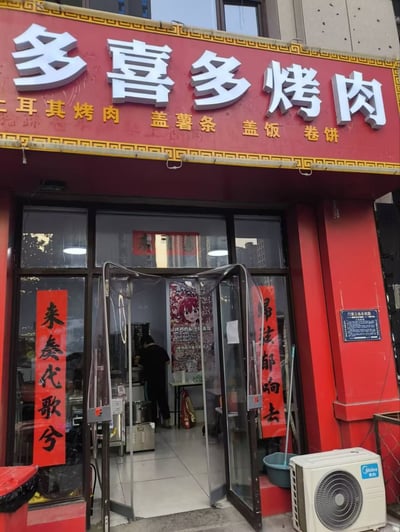 一张烤肉店的门面实拍图，店铺招牌为红色底配黄色边框，上方醒目白色大字写着“多喜多烤肉”，下方黄色小字标注经营品类“土耳其烤肉 盖薯条 盖饭 卷饼”。玻璃门两侧贴有红色对联，左侧为“来奏代歌兮”，右侧为“归去都响去”。店内可见一名工作人员在柜台后活动，店外右侧放置了一台白色美的空调和一个蓝色水桶，整体呈现出真实的街边餐饮店铺日常景象。