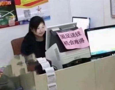 这是一张在彩票站拍摄的搞笑照片，前台电脑上贴着粉色标语写着“国足送钱 机会难得”，用来调侃中国足球队（国足）常常输球，引申为买彩票押对手稳赢，是黑色幽默和自嘲的网络段子。大量小票突出彩票站氛围。这种标语在彩民和球迷圈子广为流传，自带讽刺意味。