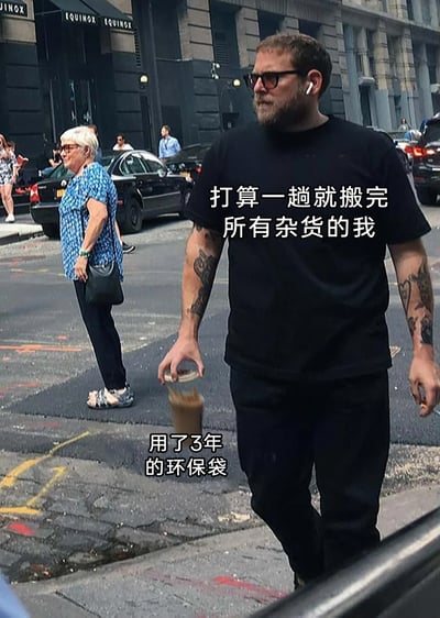 这是一张利用街头实拍的视觉错位效果制作的搞笑梗图。画面中，前方穿着黑衣、有纹身的男子的手，恰好与后方过马路的白发老人的身体位置重合，形成了“男子提着老人”的有趣错觉。图中添加的文字“打算一趟就搬完所有杂货的我”对应男子，“用了3年的环保袋”对应老人，将试图一次性搬完所有杂货的自己比作逞强的男子，而把用了3年、已经不堪重负的旧环保袋比作被“提着”的老人，生动又幽默地还原了生活中想一次性搬完所有东西，却因袋子老旧而担心它撑不住的场景，极易引发有类似经历的人的共鸣。