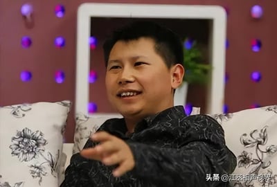 这张图片展示了相声演员何云伟参与访谈节目的场景，他坐在带有花纹靠垫的沙发上，身着深色带花纹上衣，面带笑意，嘴巴微张，同时伸出一只手做出手势，看起来正在投入地表达自己的观点。背景是带有紫色光点装饰的墙面，还有白色边框的区域，整体氛围轻松闲适，呈现出访谈聊天时的自然状态。