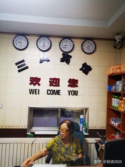 这是一张宾馆前台的实拍场景图，墙面上方悬挂着四个标注了东京、巴黎、纽约、伦敦的世界时钟，对应不同时区时间；墙面上设置了立体字标识“三A宾馆”，下方搭配红色的“欢迎您”字样，以及错误的中式英语翻译“WEI COME YOU”（正确应为WELCOME），形成趣味的语言错误笑点。前台区域有一位佩戴黄框眼镜的工作人员，正坐在椅子上操作，手中持有小本子；右侧货架摆放着饮品和杂物，墙面右上角装有监控摄像头，整体是日常的宾馆前台工作场景。
