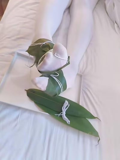 这是一张充满节日趣味的视觉错位摆拍图，画面中穿着白丝袜的双脚被粽叶和棉线以包粽子的方式捆绑起来，借助特定拍摄角度，营造出双脚化身‘巨型粽子’的搞笑视觉效果，背景是柔软的床单，整体氛围轻松诙谐，紧扣端午节包粽子的习俗玩出创意整活。
这张图利用视觉错觉制造出食物与身体部位的混淆，是节日相关的趣味脑洞创作。