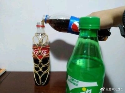 这是一张网友拍摄的趣味整活静态图，画面呈现拟人化的搞笑场景：棕色桌面上，一个可口可乐瓶被麻绳以捆绑的方式缠住瓶身直立放置，画面右上方一只手握着百事可乐瓶，正在往被绑的可口可乐瓶口倒液体，画面右前景放置着一瓶雪碧，遮挡住部分视线，仿佛在旁观整个过程。该创作利用可口可乐和百事可乐是竞品的大众认知，把饮料瓶拟人化模拟出拷问、霸凌的场面，笑点十足。
