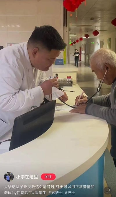 医院走廊内，一位穿着白大褂的医生身体前倾，手持纸巾靠近听诊器，对面头发花白的老人正将听诊器一端放在医生胸前，另一端塞入耳中，同时执笔在病历本上书写。背景中悬挂着红色灯笼，营造出温馨氛围，远处还有医护人员走动。画面展现了医患间暖心的互动场景，颠覆了传统医生为患者检查的模式，充满温情。