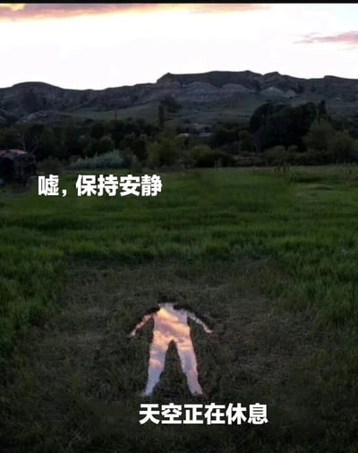 这张图片展示了一片广阔的绿色草地，远处有连绵的山脉，天空呈现出日落时分的渐变色彩。草地上有一个人形的凹陷区域，里面倒映着天空的景象，形成了独特的视觉效果。图片上方有文字'嘘，保持安静'，下方有文字'天空正在休息'，整体营造出一种宁静、诗意的氛围。