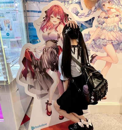 A photograph showing a person with long black pigtails wearing a white blouse and black skirt, posing with their hand on the head of a large anime character cutout. The cutout features a pink-haired female character in a purple and black dress. In the background, another anime character cutout with blue hair wearing a white and purple dress is visible. The setting appears to be an anime or entertainment venue with glass windows and various signs visible in the background. The 'hololive' logo is visible at the bottom of the cutout, indicating these characters are likely from the Hololive virtual YouTuber agency.