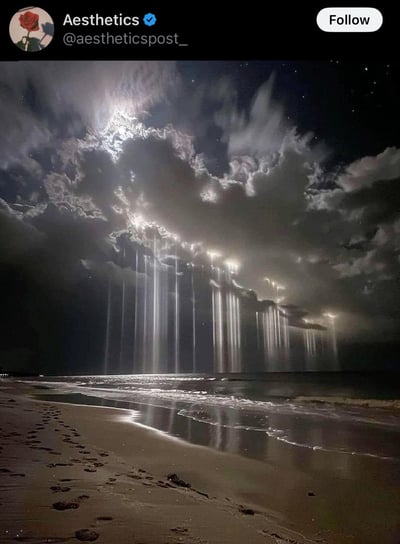 这是一幅AI生成的奇幻夜景图，画面中夜幕下的海滩静谧而梦幻：厚重的夜空云层被光源照亮边缘，一道道如同瀑布般的光束从云层间倾泻而下，在海面上形成粼粼光影，沙滩上的一串脚印为这个超现实场景增添了一丝真实感，整体氛围震撼又治愈，仿佛是异世界的海岸奇景。