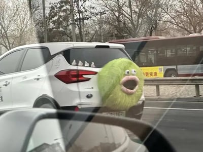 这张图片是一辆白色SUV行驶在路上，通过P图方式将汽车尾部合成了一个大嘴绿色毛毛球怪兽头像，搞怪且充满创意。这个怪兽的表情夸张、嘴巴大开，看起来像是在惊叫，引发强烈视觉反差和幽默感。图片属于错位梗和恶搞风格，适合用来表达震惊或“看呆了”。未发现谐音梗，但有明显视觉幽默。