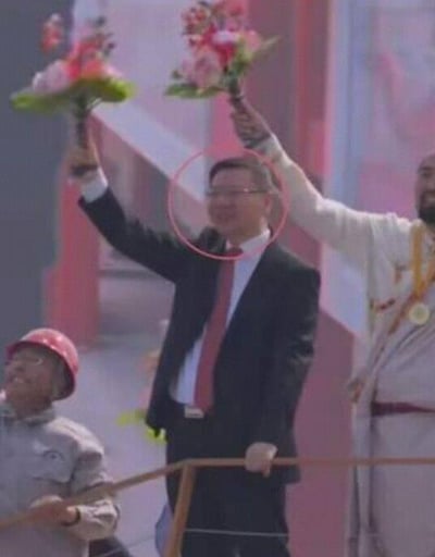 Three people appear to be at a celebratory event or ceremony. The central figure is wearing a black suit with a red tie and holding up a bouquet of pink and white flowers. His face has been circled in red, drawing attention to him. To his right, another person in white traditional-style clothing is also holding up flowers. To his left, a person wearing a red hard hat is looking upward. The background appears to be an indoor stage with red and white colors.