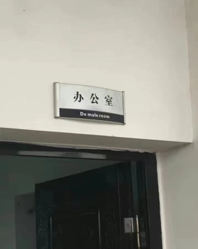 这是一张拍摄于室内门口的实拍照片，门上方的墙面上固定着一块带有金属边框的门牌。门牌上半部分的白色区域印有黑色简体中文“办公室”，而下半部分的黑色条块区域内，却错误地标注了英文“Do male room”，该英文表述完全不符合“办公室”的正确英文翻译，属于明显的翻译失误，推测是使用不准确的翻译工具或工作人员失误导致的乌龙情况，让原本普通的门牌变得滑稽好笑。