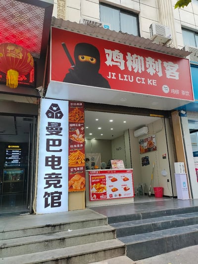 这是一张线下商铺实拍图，画面主体是名为「鸡柳刺客」的小吃店：红色招牌上印有持剑的卡通刺客形象，标注了店名拼音及「鸡柳 茶饮」的经营品类；门店入口处的立牌展示了香酥鸡柳、招牌三合一、吮指薯条、空气年糕等主打餐品，店内可见价目表与餐品海报，还有工作人员在店内。门店左侧是「曼巴电竞馆」的发光灯牌，旁边还有其他商铺的红色灯笼，整体呈现日常的街边商业街场景。
店名「鸡柳刺客」玩梗自网络热词「雪糕刺客」，暗示该店鸡柳可能价格超出预期。