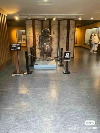 这是一张在博物馆展厅内拍摄的静态照片，画面核心是一尊具有生殖崇拜文化意义的深色雕像，雕像呈蹲坐姿态，造型突出展示男性生殖器官，被黑色隔离栏与绳索围起保护。雕像旁设有“免费讲解等候点”的标识牌和“请勿触摸”的提示牌。背景是绘有传统人物图案的折叠屏风，展厅墙面为暖黄色调，地面铺设灰色纹理瓷砖，画面右侧有两位身着休闲装的参观者正在观看其他展柜内的展品。整体空间氛围庄重，是典型的文博场馆展陈场景，用于向观众展示民俗文化相关的特色文物。