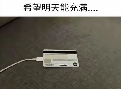 这是一张搞笑梗图，采用顶部配文加下方实拍图的形式，顶部文字为“希望明天能充满....”，下方实拍内容是一根充电线的插头插在平放在灰色桌面的银行卡磁条位置，假装正在给银行卡充电，用违背常识的荒诞操作，调侃发布者希望银行卡余额增加、收入上涨的诉求，笑点在于将电子设备充电的概念迁移到银行卡上，暗指想要给余额“充值”的愿望。