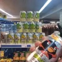 在超市的货架区域，印有《愤怒的小鸟》绿猪形象的饮料罐被创意堆叠成了绿猪的立体造型，拍摄者手持印有黑鸟形象的同款联名饮料，完美还原游戏里黑鸟与绿猪对阵的经典设定，充满了趣味性和创意感。