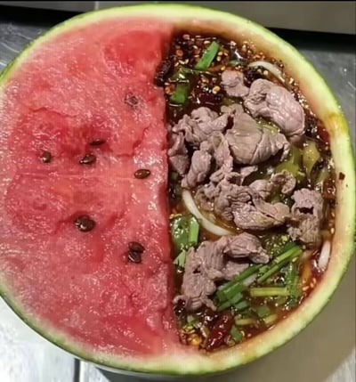 这张静态图片展示了被横向切开的半颗西瓜，西瓜内部被分为两个泾渭分明的区域：左侧保留了原生的红色西瓜果肉，上面散落着数颗黑色西瓜籽；右侧的西瓜瓤被全部挖空，盛放了一份麻辣风味的冒菜，冒菜中包含牛肉片、葱段、香菜、干辣椒、芝麻、粉条等食材，浸在红亮的麻辣汤汁中。两种口味、属性完全不同的食物被放置在同一个西瓜壳容器中，视觉反差强烈，属于创意新奇的黑暗料理。